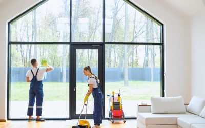 Young cleaners in blue uniform mopping wooden floor, cleaning big panoramic window, preparing country house for selling. Successful cleaning service concept